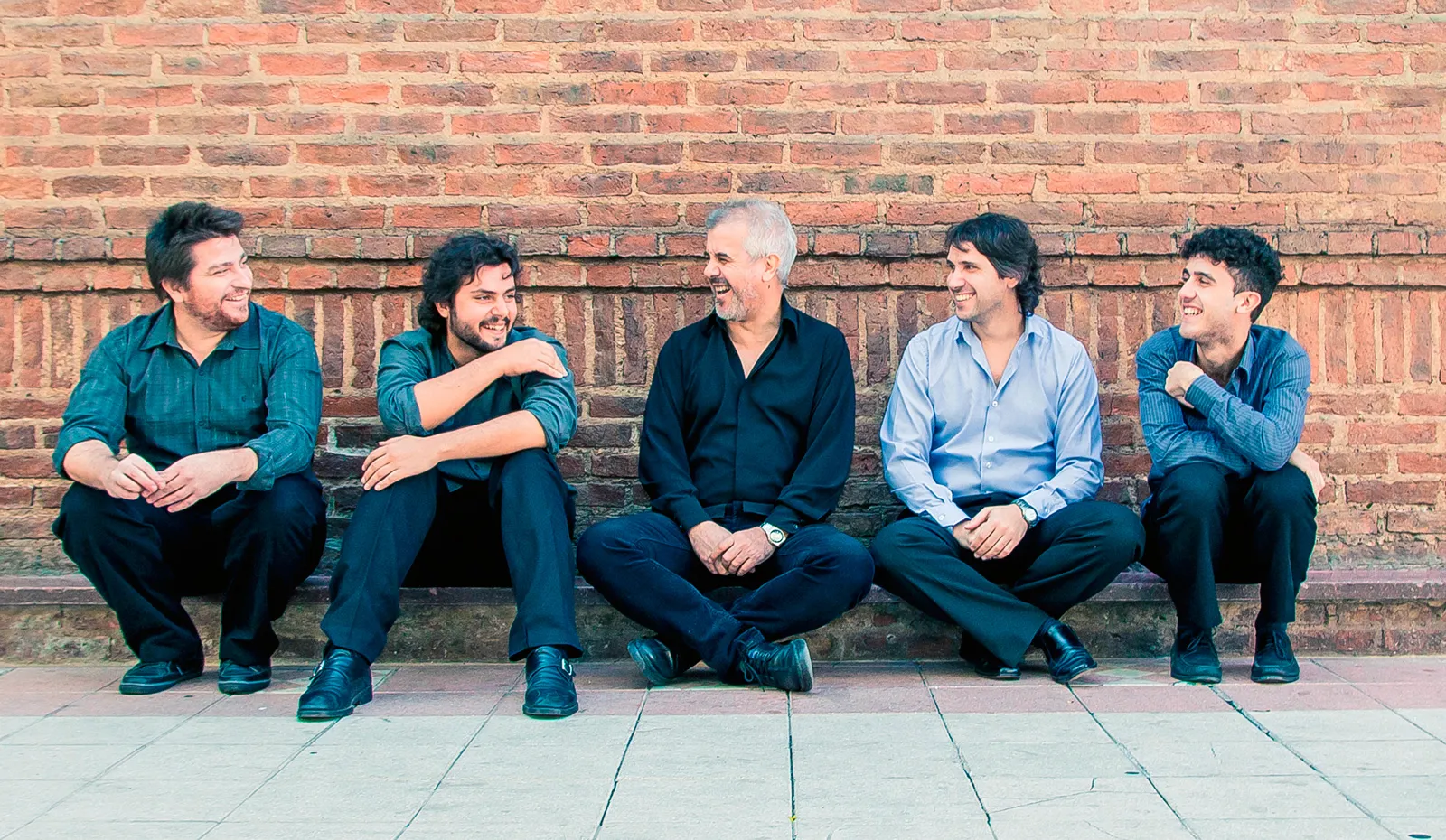 Roberto Calvo y otros cuatro músicos sentados contra una pared de ladrillo, riéndose